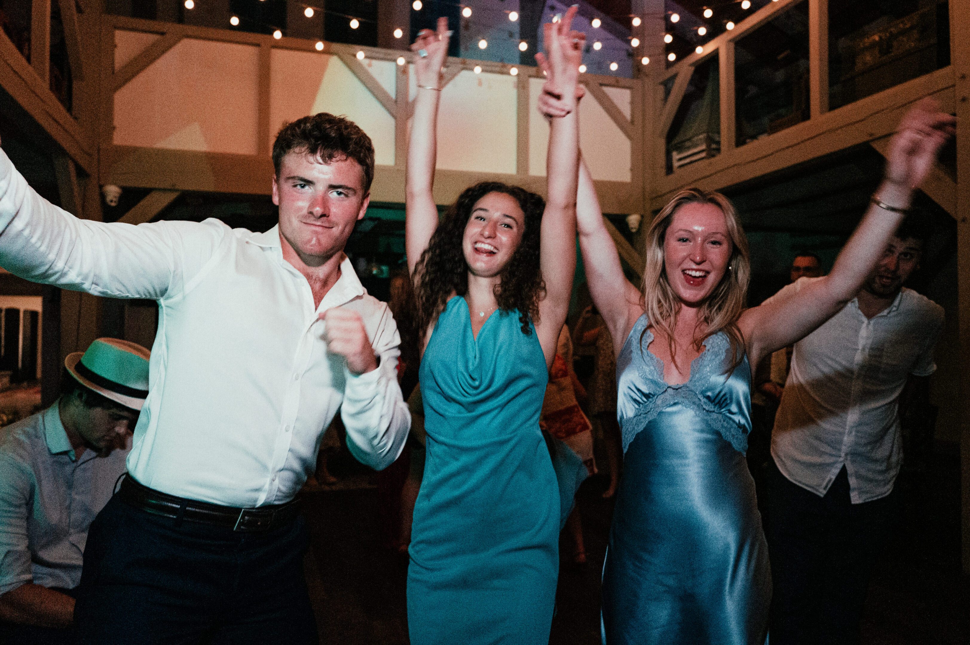 Three friends dancing and celebrating indoors under string lights.